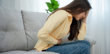 A woman sitting on a couch, holding her stomach as if she is experiencing stomach pain.